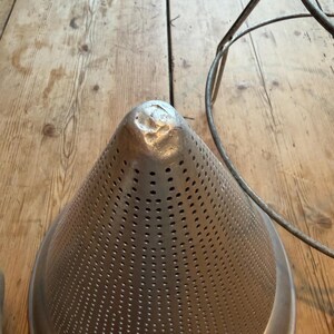 May include: A conical metal food mill or strainer featuring a perforated surface with numerous small holes and a solid, rounded tip. The object is constructed from a silver-toned metal and is positioned next to a thin, circular metal wire frame.