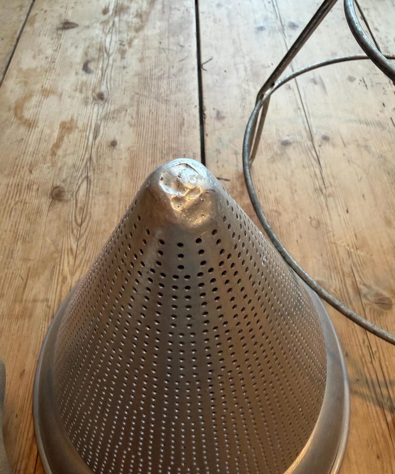 May include: A conical metal food mill or strainer featuring a perforated surface with numerous small holes and a solid, rounded tip. The object is constructed from a silver-toned metal and is positioned next to a thin, circular metal wire frame.