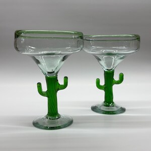 May include: Two clear glass margarita glasses featuring stems shaped like green cacti with small arms. The bowls of the glasses are wide and circular with a subtle green tint along the rim, supported by a solid green glass base and stem.