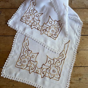 May include: White linen table runner and matching napkin with embroidered floral designs in shades of brown. The edges of both pieces are trimmed with a delicate white lace.