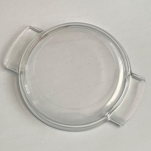 May include: A clear, round glass lid with two curved handles. The lid is designed to fit a round dish and is made of transparent glass. The handles are integrated into the design, providing a secure grip.
