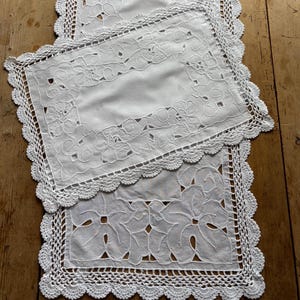 May include: White embroidered table runners and placemats with a floral design. The placemats and runners have a scalloped crochet lace edge. The table runners and placemats are on a wooden surface.