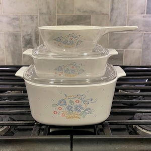 May include: Three white ceramic casserole dishes with floral designs and clear glass lids. The dishes are stacked on top of each other on a black stovetop.