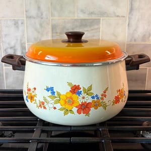May include: A vintage white enamel pot with a vibrant floral design in yellow, orange, red, and blue. The lid is gradient orange to yellow, and the handles are brown. This cooking pot is a retro kitchenware item.
