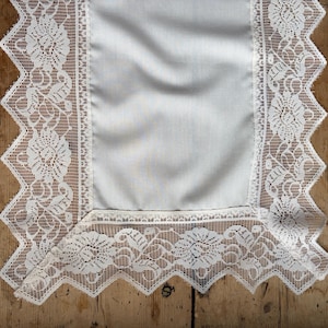 May include: A white lace table runner with a scalloped edge and floral patterns. The center is a solid white fabric. The runner is placed on a wooden surface, creating a contrast between the delicate lace and the rustic wood.