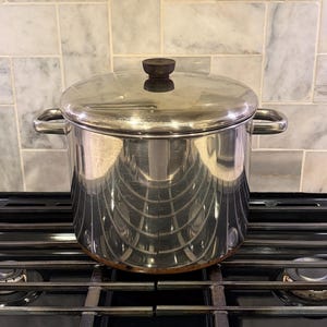 May include: A stainless steel stockpot with a lid and two side handles. The lid has a dark brown wooden knob. The pot is sitting on a black stovetop. The pot is reflective, showing the surrounding environment.