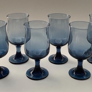 May include: Six blue-tinted stemmed glasses. Each glass has a rounded bowl and a dark blue stem and base. The glasses are arranged in a group, showcasing their design and color. The glassware is suitable for beverages.