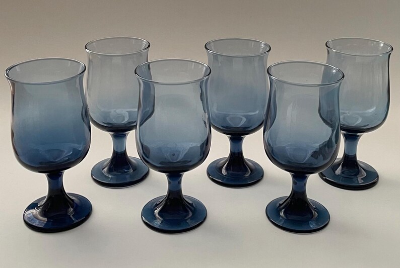 May include: Six blue-tinted stemmed glasses. Each glass has a rounded bowl and a dark blue stem and base. The glasses are arranged in a group, showcasing their design and color. The glassware is suitable for beverages.