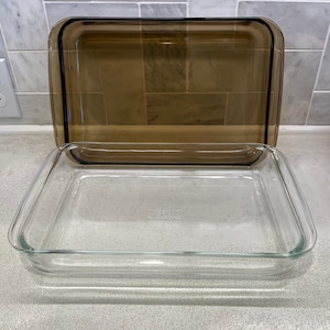 May include: A clear glass rectangular baking dish with a brown tinted glass lid. The dish is empty and sitting on a speckled countertop. The lid is slightly ajar, revealing the interior of the dish. The dish is designed for baking and cooking.