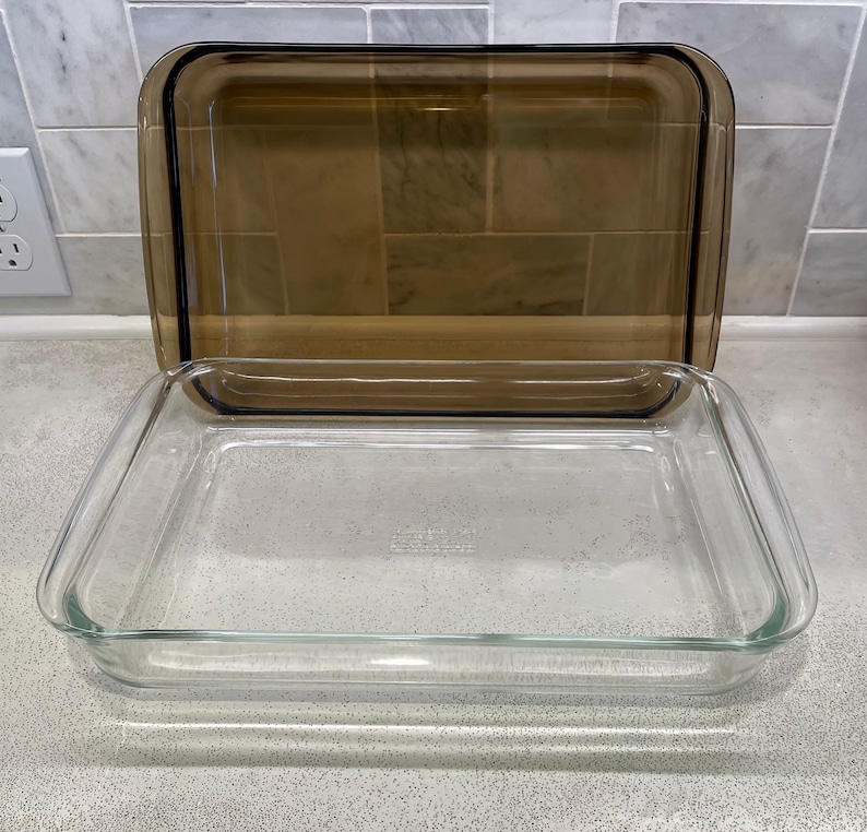 May include: A clear glass rectangular baking dish with a brown tinted glass lid. The dish is empty and sitting on a speckled countertop. The lid is slightly ajar, revealing the interior of the dish. The dish is designed for baking and cooking.