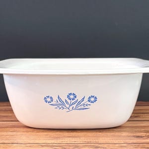 May include: A white rectangular baking dish with rounded corners and a wide rim. The exterior features a centered blue floral pattern consisting of three stylized flowers with leaves. The dish has a smooth, glossy finish and a deep basin design.