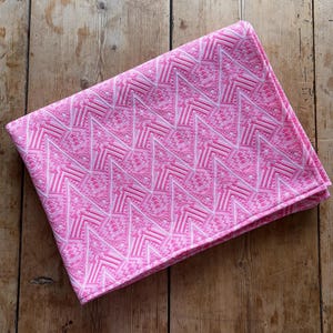 May include: Folded pink fabric with a repeating geometric pattern in white and shades of pink. The fabric is folded neatly and rests on a wooden surface.