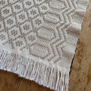 May include: A woven textile runner featuring a light beige and tan geometric pattern. The design consists of a repeating diamond grid on one side and vertical wavy lines on the other, finished with a long, straight fringe at the edge.