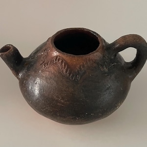 May include: A small, round, dark brown ceramic teapot with a spout and handle. The teapot has a textured surface and a wide opening at the top. The handle is a simple loop, and the spout is short and cylindrical.