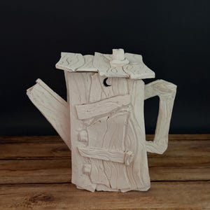 May include: A white ceramic teapot with a unique outhouse design. The teapot features a textured wooden appearance, a small crescent moon cutout, and a handle. The spout and lid are also visible. The teapot is set against a dark background.