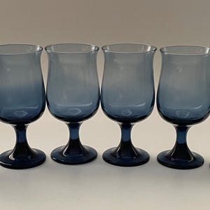 May include: Six blue-tinted stemmed glasses arranged in a row. The glasses have a tulip-shaped bowl and a dark blue base and stem. The color of the glass fades from a lighter blue at the top to a darker blue at the base.