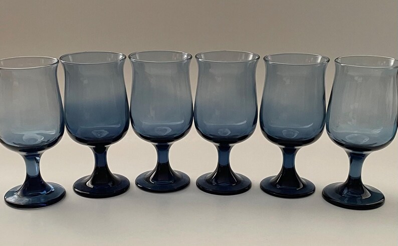 May include: Six blue-tinted stemmed glasses arranged in a row. The glasses have a tulip-shaped bowl and a dark blue base and stem. The color of the glass fades from a lighter blue at the top to a darker blue at the base.