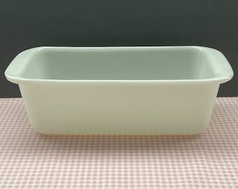 May include: A rectangular, off-white ceramic baking dish with rounded handles. The dish is empty and sits on a pink and white checkered surface. The background is a solid dark gray.