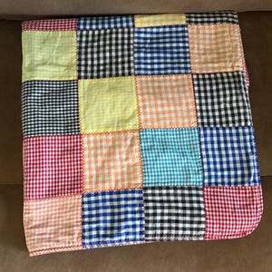 May include: A patchwork quilt featuring squares of various colors and gingham patterns. The quilt includes red, black, blue, yellow, and orange squares, all stitched together with a contrasting thread. The quilt is folded and appears to be made of cotton.