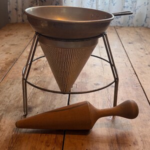 May include: A metal food mill or chinois strainer with a conical perforated body, resting on a three-legged wire stand. The set includes a separate, smooth, light brown wooden pestle with a tapered conical end and a rounded handle for pressing.