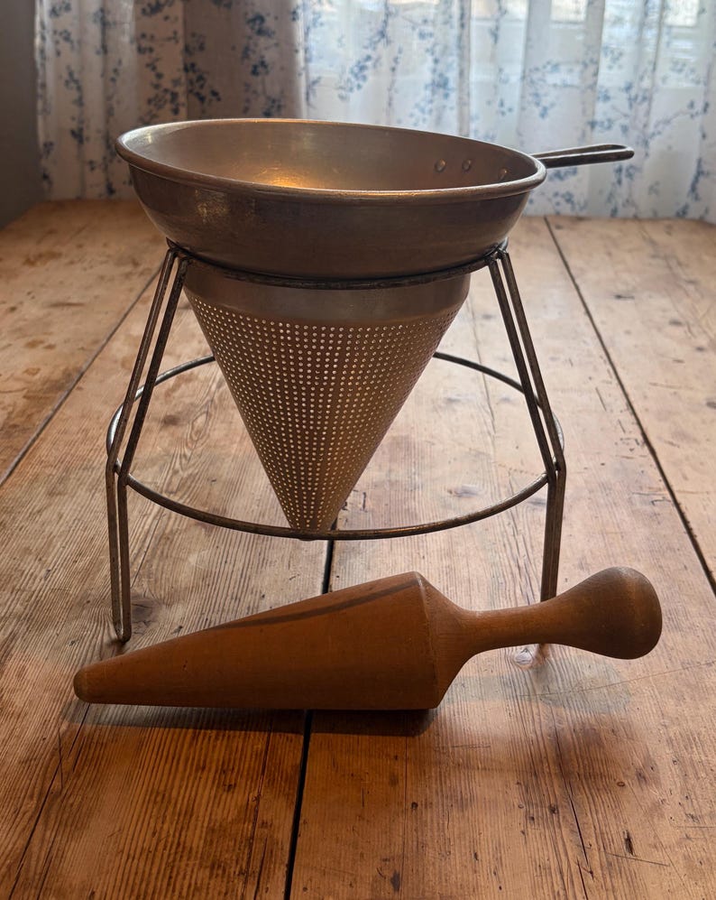May include: A metal food mill or chinois strainer with a conical perforated body, resting on a three-legged wire stand. The set includes a separate, smooth, light brown wooden pestle with a tapered conical end and a rounded handle for pressing.