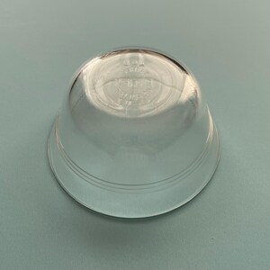 May include: A clear, glass bowl with a rounded shape and a wide, flat base. The bowl has a slightly flared rim and three concentric rings near the base. The bottom of the bowl has etched text. The bowl is sitting on a light blue surface.