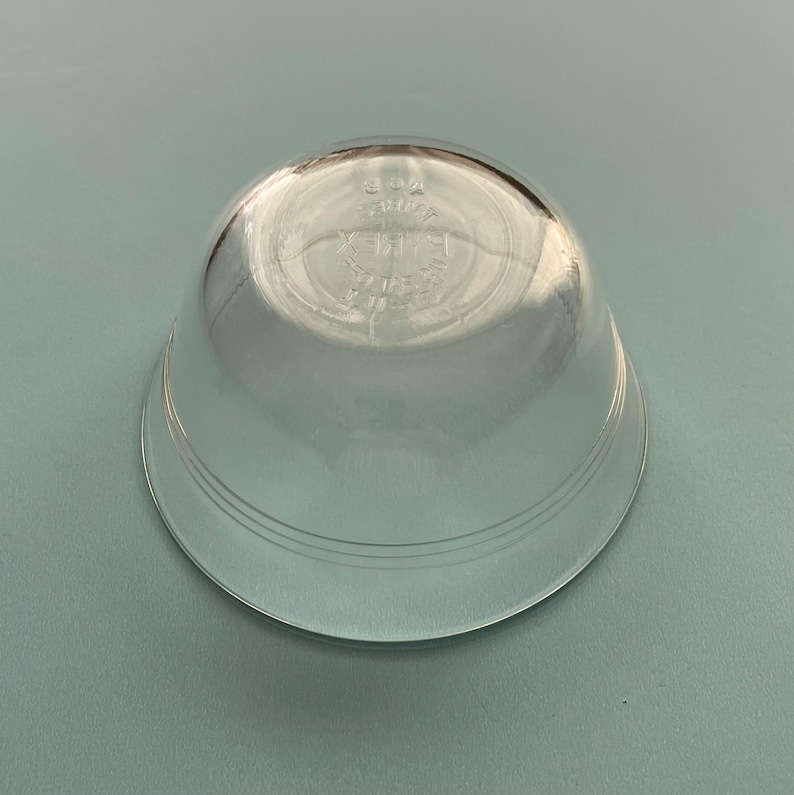 May include: A clear, glass bowl with a rounded shape and a wide, flat base. The bowl has a slightly flared rim and three concentric rings near the base. The bottom of the bowl has etched text. The bowl is sitting on a light blue surface.