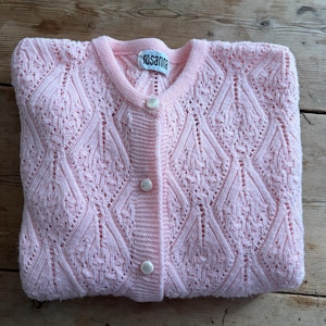 May include: A light pink knitted cardigan with a round neckline and three white buttons. The cardigan features a detailed openwork pattern and a label that reads "rosanna". The garment is folded on a wooden surface.