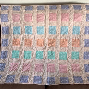 May include: A handmade quilt with a patchwork design. The quilt features squares in pastel colors: pink, blue, orange, and green, each with a white polka dot pattern. The squares are set against a neutral background.