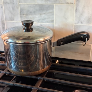 May include: A stainless steel saucepan with a copper-colored base, featuring a polished metal lid with a black knob. The pan has a long, black heat-resistant handle with a small metal hanging loop at the end, reflecting the surrounding kitchen environment.