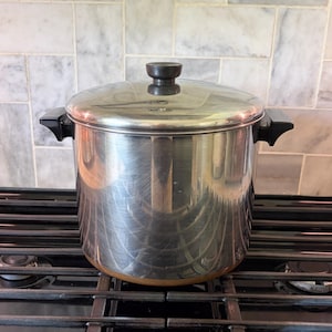 May include: A large stainless steel stockpot with a polished finish, featuring two black side handles and a matching stainless steel lid with a black knob. The pot has a cylindrical body and a flat base, designed for use on a stovetop surface.