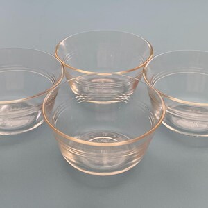 May include: Four clear glass bowls with a slightly flared rim. Each bowl has a rounded shape and a subtle, horizontal ridge detail near the base. The bowls are arranged on a light blue surface.