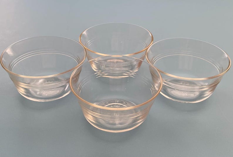 May include: Four clear glass bowls with a slightly flared rim. Each bowl has a rounded shape and a subtle, horizontal ridge detail near the base. The bowls are arranged on a light blue surface.