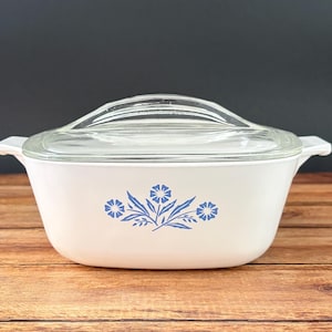 May include: A white rectangular casserole dish with integrated side handles and a clear glass lid. The front of the dish features a blue floral pattern consisting of three stylized flowers with leaves centered on the smooth white surface.