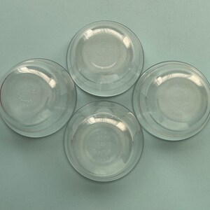 May include: Four clear glass bowls arranged on a light blue surface. The bowls are round with a slightly flared rim and a clear, transparent appearance. Each bowl has a circular design in the center.
