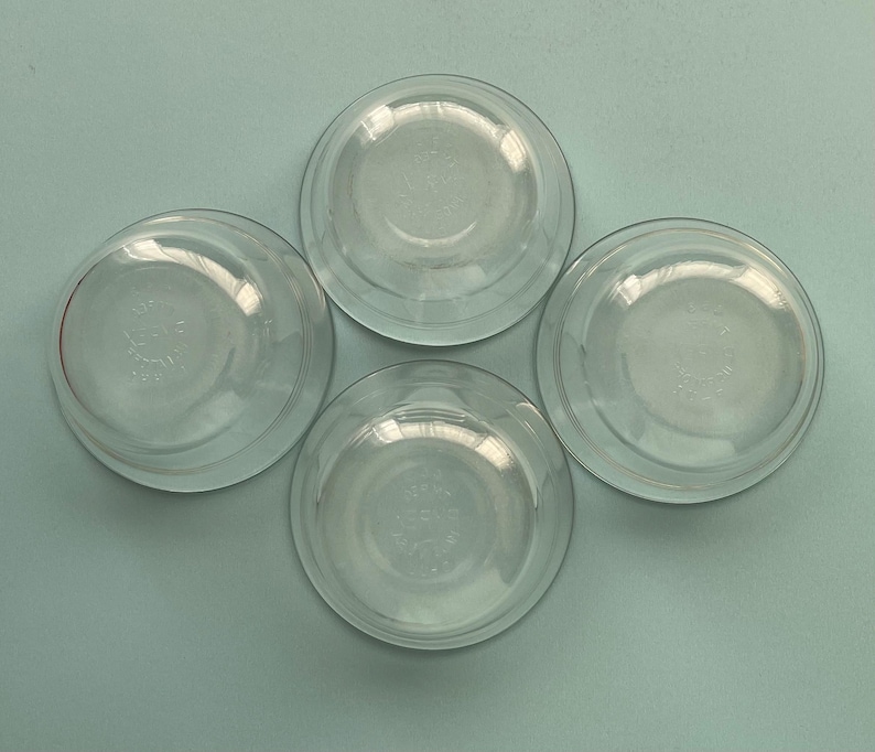 May include: Four clear glass bowls arranged on a light blue surface. The bowls are round with a slightly flared rim and a clear, transparent appearance. Each bowl has a circular design in the center.