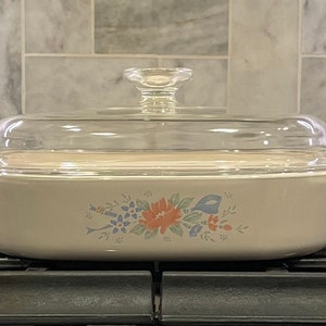 May include: An off-white rectangular ceramic casserole dish with integrated side handles and a clear glass lid featuring a central knob. The front of the dish displays a floral pattern in muted shades of orange, blue, and green.
