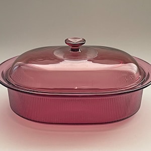 May include: A vintage, oval-shaped, ruby-colored glass casserole dish with a matching lid. The dish has a ribbed exterior and a clear glass lid with a small knob handle. The dish is designed for baking and serving.