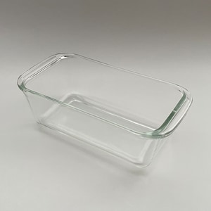 May include: A clear, rectangular glass loaf pan with rounded corners. The baking dish is empty and sits on a white surface. The pan is designed for baking bread, cakes, or other baked goods.
