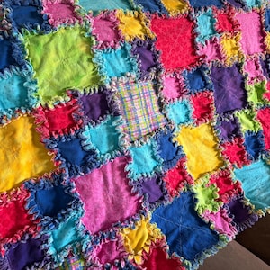 May include: A colorful patchwork quilt featuring squares of various colors including pink, blue, yellow, green, and purple. The edges of each square are frayed, creating a textured effect. The quilt is made of soft fabric.