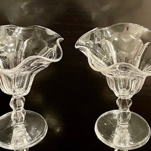 May include: Two clear glass dessert dishes with a ruffled rim and a pedestal base. The dishes have a delicate, elegant design, perfect for serving ice cream or other treats. The glass is transparent, allowing the contents to be fully visible.