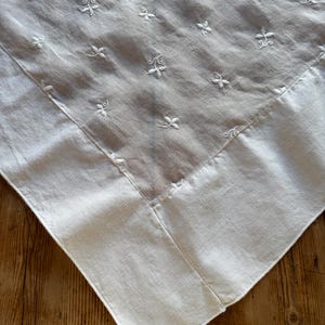May include: A white, square tablecloth with delicate floral embroidery. The embroidery is evenly spaced across the fabric, and the edges are neatly hemmed. The tablecloth is made of a soft, light-colored fabric.