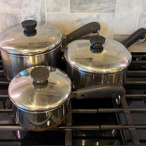 May include: A set of three stainless steel pots with matching lids, each featuring a black handle and a black knob on the lid. The pots vary in size and are arranged on a black stovetop grate, showcasing a polished metallic finish throughout.