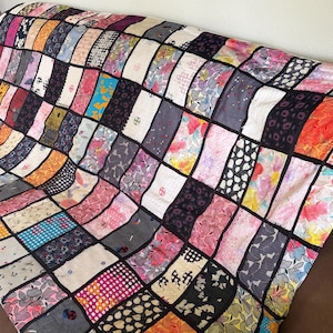 May include: A colorful patchwork quilt with a variety of patterned fabrics in a grid-like design. The quilt features a mix of floral, polka dot, and geometric patterns in shades of pink, blue, orange, and black. The quilt is draped over a brown surface.