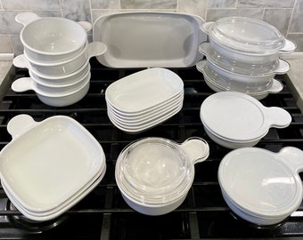 May include: A set of white ceramic dishes with lids and handles. The dishes are arranged on a black stovetop.