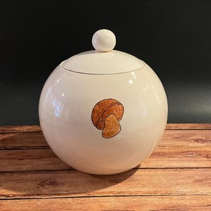 May include: A white, spherical ceramic jar with a lid and a small, round knob. The jar features a hand-painted brown mushroom illustration on the front. The jar is sitting on a wooden surface.