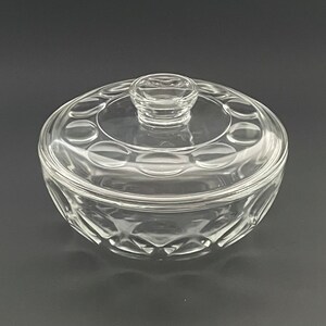 May include: A clear glass serving dish with a lid. The dish is round with a decorative pattern of circles around the rim and on the lid. The lid has a small knob for easy handling. The dish is designed for serving food.