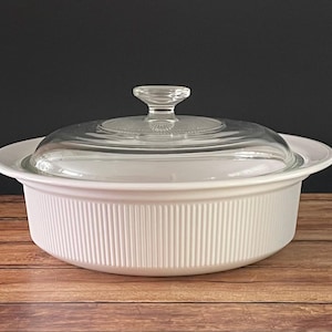 May include: A white, ribbed casserole dish with a clear glass lid. The dish sits on a wooden surface against a dark background. The lid has a small handle. This kitchenware is ideal for baking and serving.