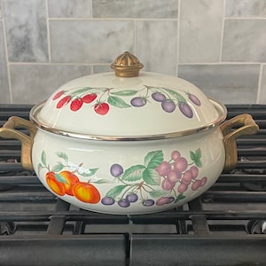 May include: Cream-colored enamelware pot with lid, adorned with fruit illustrations. The pot features a gold-colored handle and lid knob. The fruit designs include red strawberries, purple plums, and orange pumpkins. The pot is sitting on a black stove.