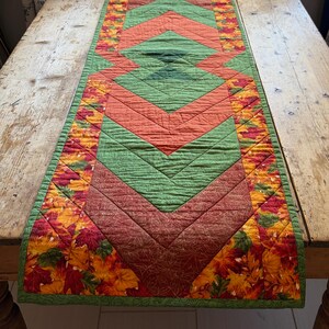 May include: A rectangular quilted table runner featuring a central geometric chevron pattern in shades of green, orange, and brown. The border is decorated with a vibrant print of autumn leaves in red, yellow, and orange, finished with green trim.